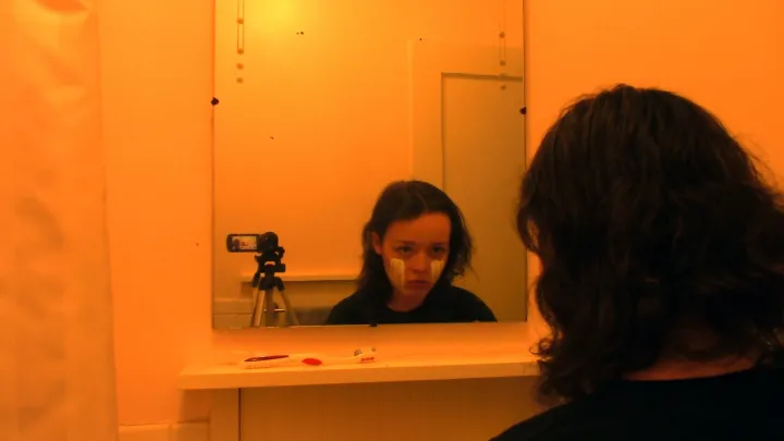 A teenager looks into a mirror. A camera watches her watching herself. 