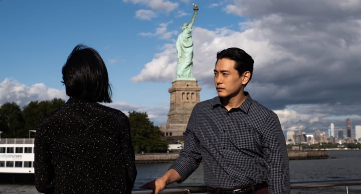 Greta Lee (turned away from us) looks at the Statue of Liberty. Teo Yoo (turned toward us) looks away.